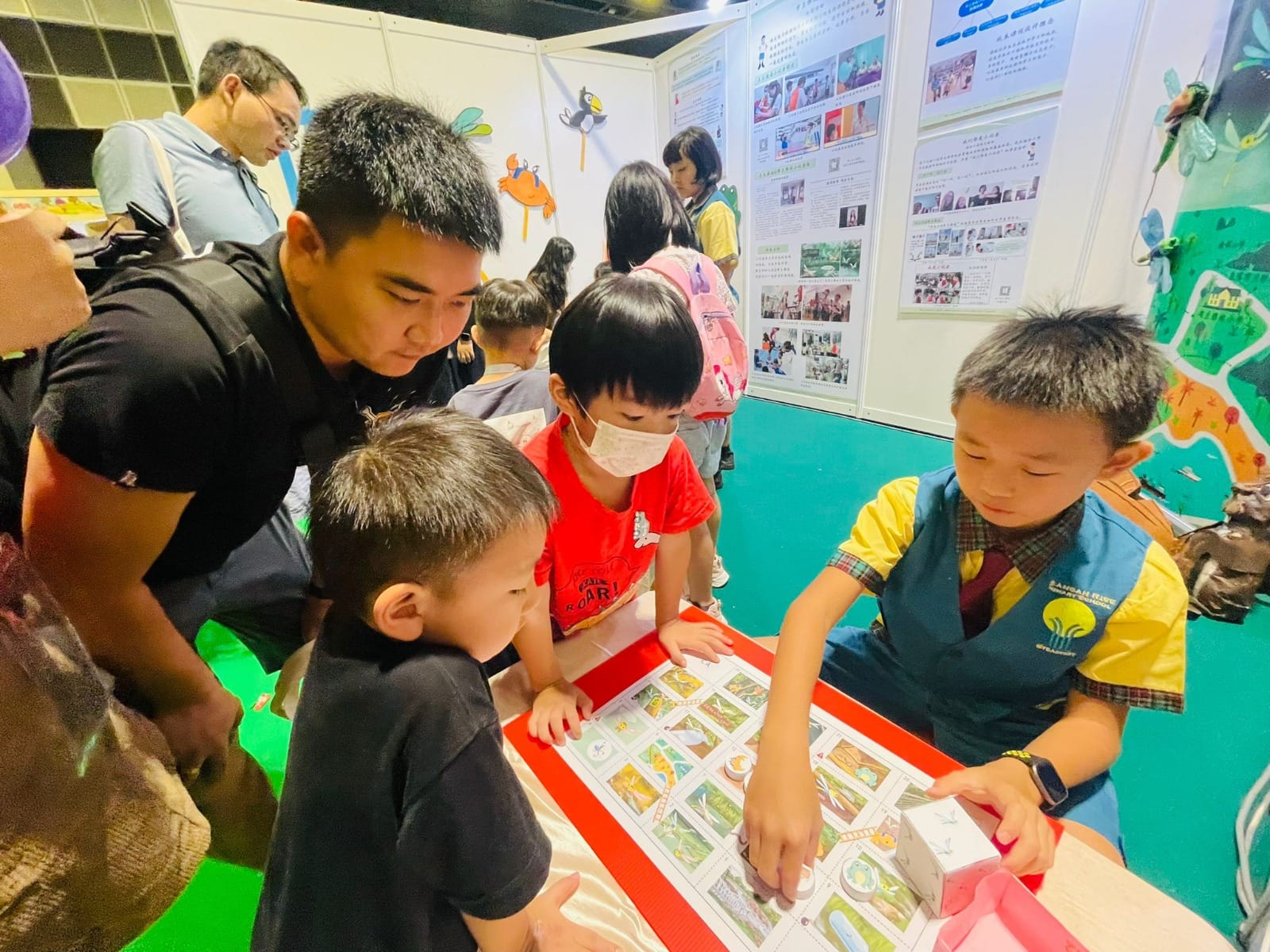 An adult man and three young children look at illustrated cards on a table at an indoor educational exhibition.